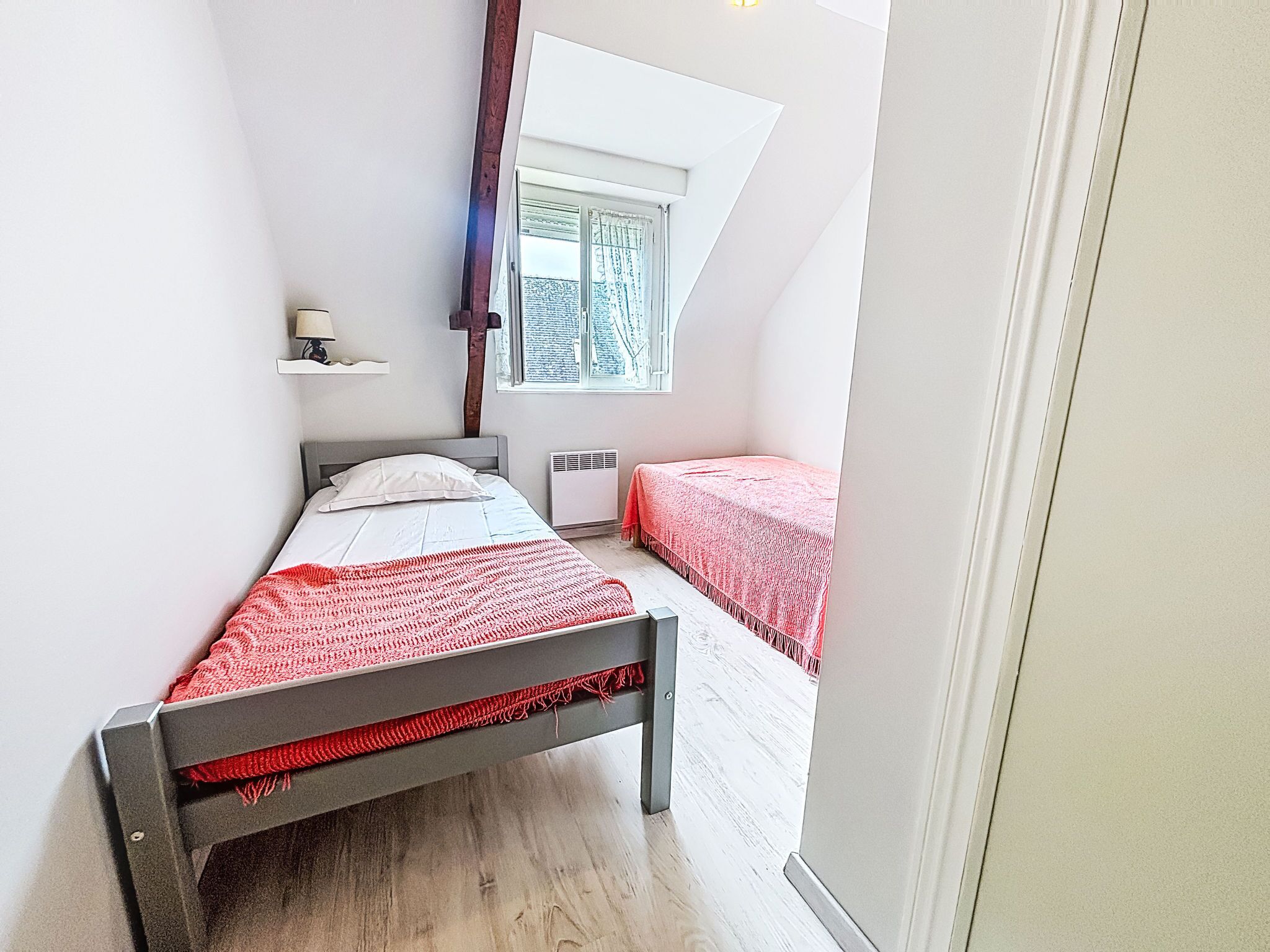 A bedroom with a sloped ceiling, a bed with a red blanket, a bedside table, a window with curtains, and a radiator.
