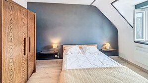 A bedroom with a sloped ceiling, a bed with white linens, two bedside lamps, a wooden wardrobe, and a large window. - Maison Le Magouër by Interhome (Plouhinec Morbihan)