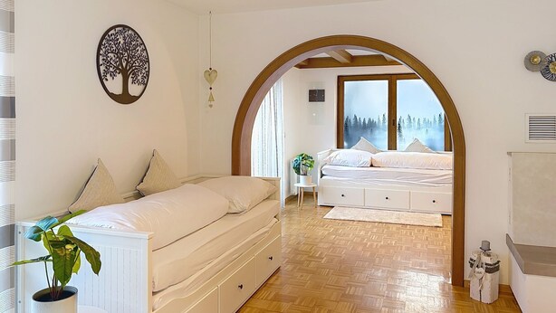 A bedroom with a bed, a bedside table with a plant, a wooden floor, and a large window with a view of trees. - Sommer Apartements Schwarzwald by Interhome (Simonswald)
