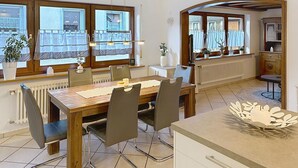 A dining area with a wooden table and chairs, large windows with curtains, a radiator, and a kitchen area with cabinets and a countertop. - Sommer Apartements Schwarzwald by Interhome (Simonswald)