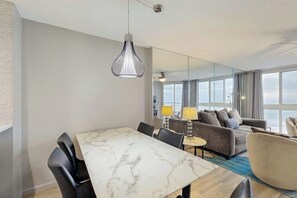 A modern hotel room with a marble dining table, black chairs, a grey sofa, a round armchair, and a large window offering a view of the sea. - Oceanfront condo with private balcony, indoor pool & direct beach access (North Myrtle Beach)
