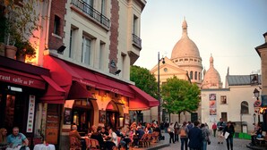 Eine Kopfsteinpflasterstraße mit Außenbestuhlung eines Restaurants, ein Kuppelgebäude im Hintergrund und ein klarer Himmel.
