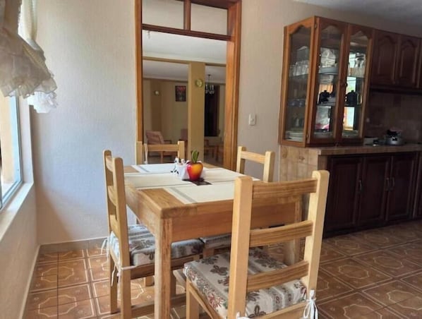A dining area with a wooden table and chairs, a fruit on the table, and a kitchen with cabinets and a window.