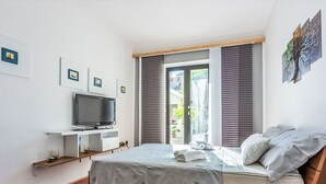 Un dormitorio con cama, televisor sobre un mueble, ventana con persianas y obras de arte en las paredes.
