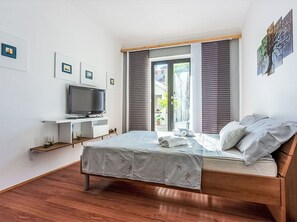Ein Schlafzimmer mit Bett, Fernseher auf einem Schrank, einem Fenster mit Jalousien und Bildern an den Wänden.