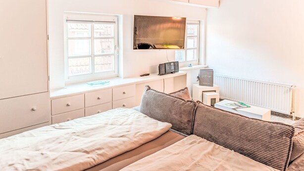 Un dormitorio con cama, televisión montada en la pared, ventana y radiador.