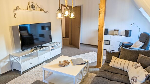 Ein modernes Wohnzimmer mit Flachbildfernseher, weißen Möbeln, Holzbalkendecke und einer gemütlichen Sitzecke.