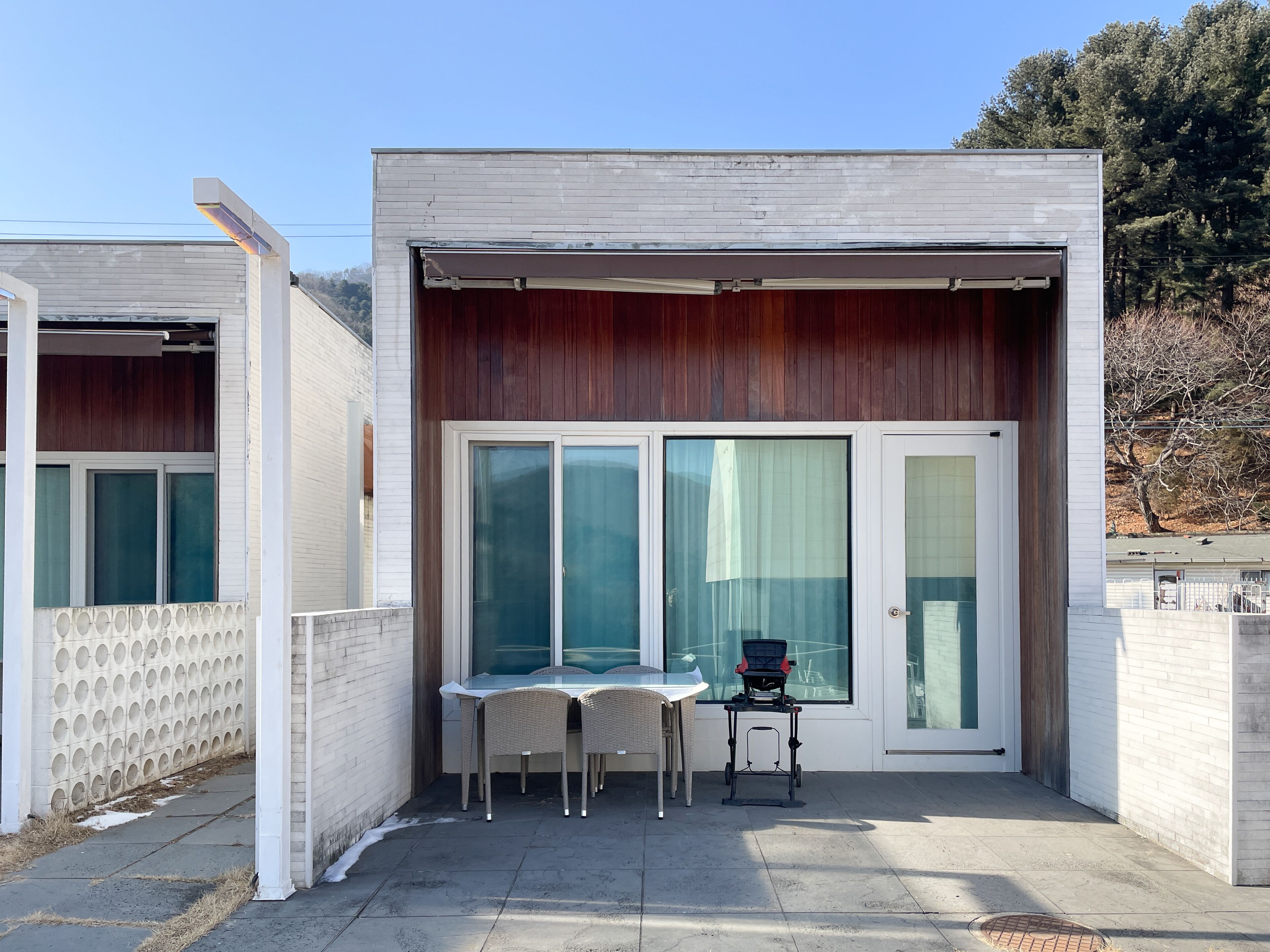 Un pequeño edificio moderno con un patio que incluye una mesa, sillas y una silla de ruedas, además de amplias puertas correderas de vidrio.
