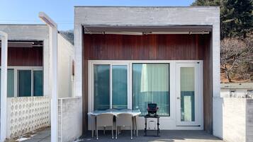 Un pequeño edificio moderno con un patio que incluye una mesa, sillas y una silla de ruedas, además de amplias puertas correderas de vidrio.