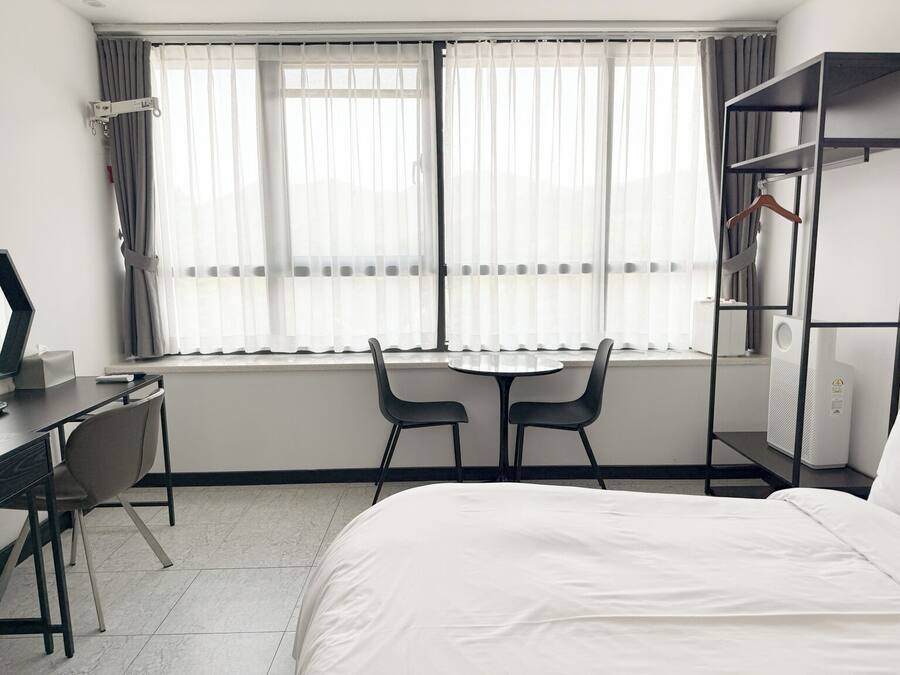 A hotel room with a bed, a desk with a computer, two chairs, a window with curtains, and a wardrobe.
