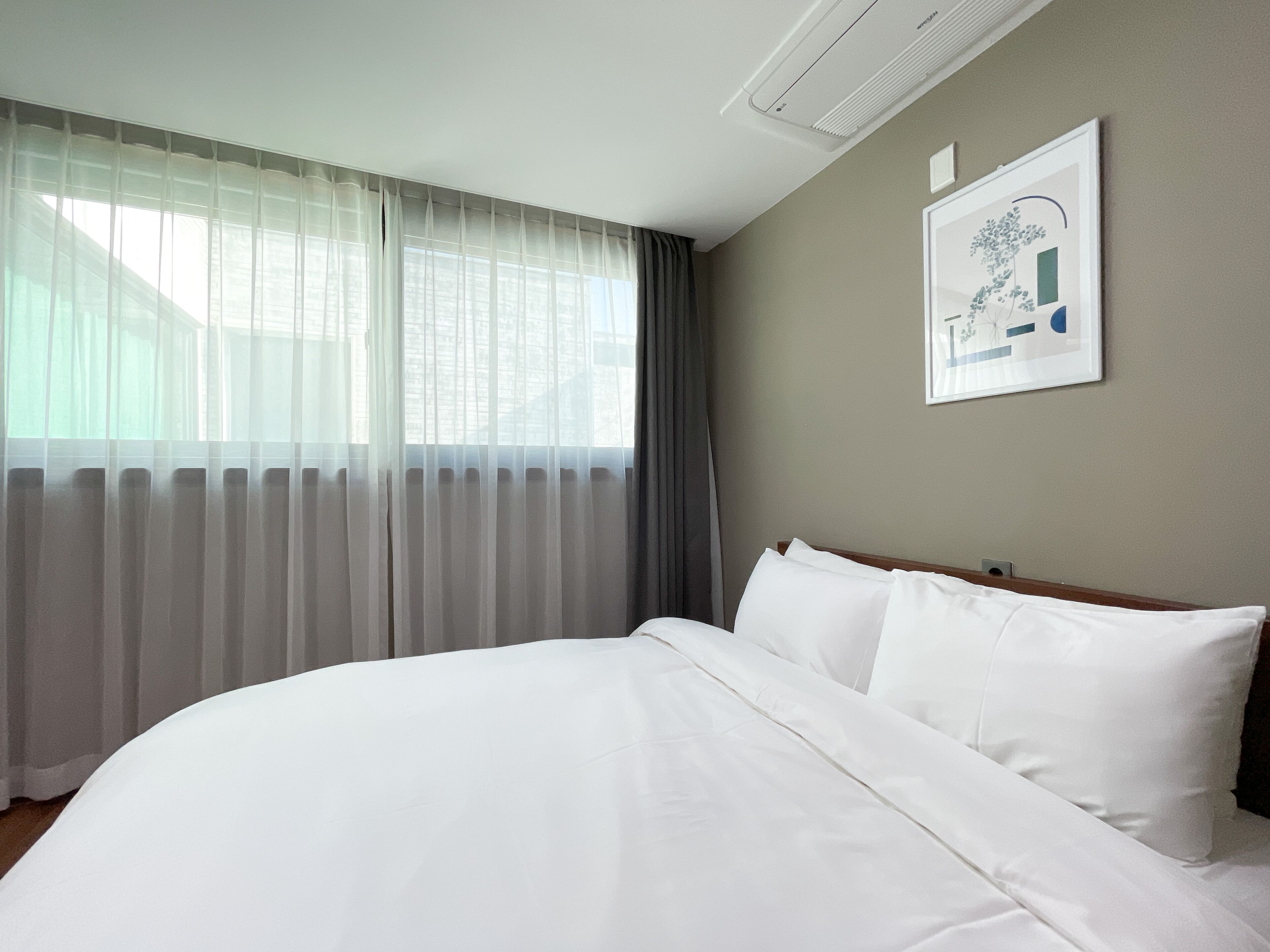 A hotel room with a large bed, a window with sheer curtains, a wall-mounted air conditioner, and a framed picture.