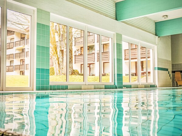 En swimmingpool med klart vand, omgivet af store vinduer med udsigt til træer og en bygning.
