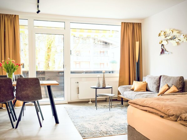 Ett modernt rum med en soffa, ett matbord med stolar, ett litet sidobord och utsikt genom stora fönster.