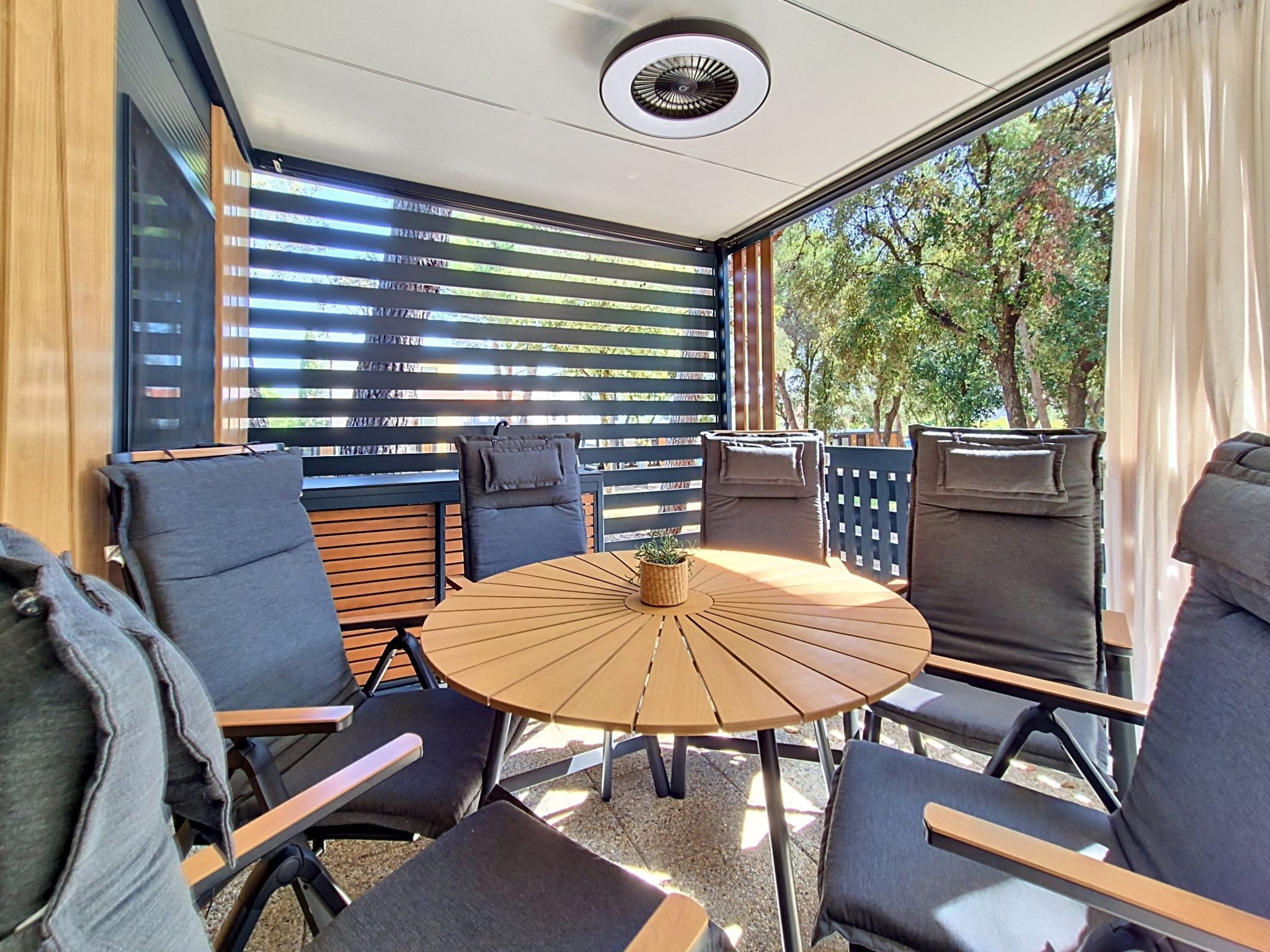 Un espace extérieur moderne, aménagé pour se détendre, avec une table ronde et des chaises, un ventilateur de plafond et une vue sur les arbres à travers de grandes fenêtres.