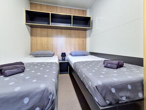 Kahden hengen huone, jossa on kaksi sänkyä, ja jokaisessa sängyssä on tyyny ja rullalle taitettu pyyhe. Sänkyjen yläpuolella on hylly ja sänkyjen välissä yöpöytä, jossa on lamppu.