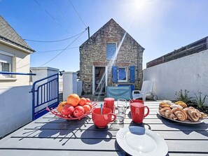 Una mesa con frutas, vasos rojos y pasteles, una silla azul y un edificio de piedra al fondo.