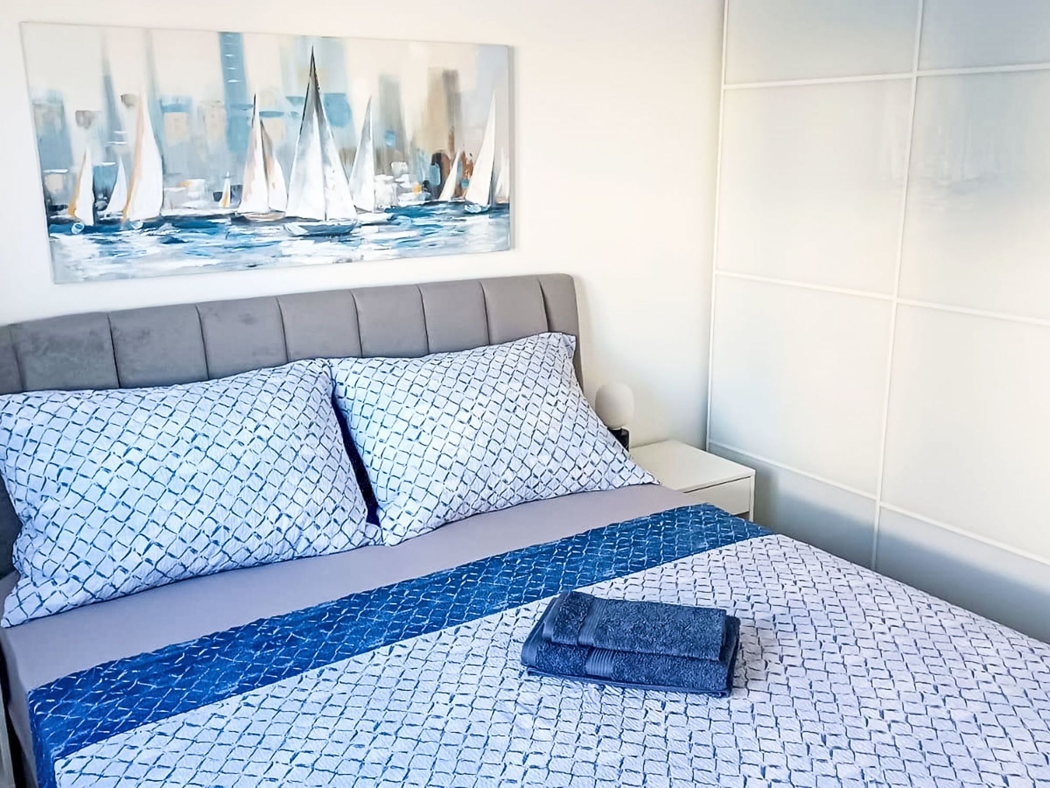 Ein Bett mit gemusterten Betttextilien, einem Kopfteil, einem Nachttisch und einem Bild von Segelbooten an der Wand.