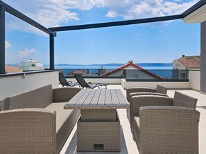 A rooftop terrace with wicker furniture, a table, and a view of the sea and distant buildings. - Sunny Terrace II by Interhome (Trogir/Okrug Gornji)