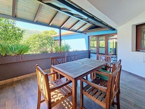 Une terrasse couverte avec une table et des chaises en bois, une vue sur la verdure et un bâtiment en arrière-plan.