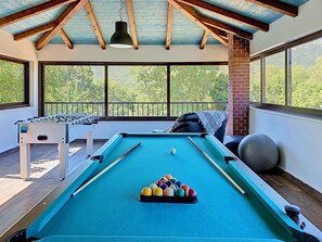 Une table de billard avec des billes, une table de baby-foot et un coin salon avec vue sur les arbres.