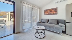 Een moderne woonkamer met een grijze bank, een ronde salontafel en een groot raam met dunne gordijnen.