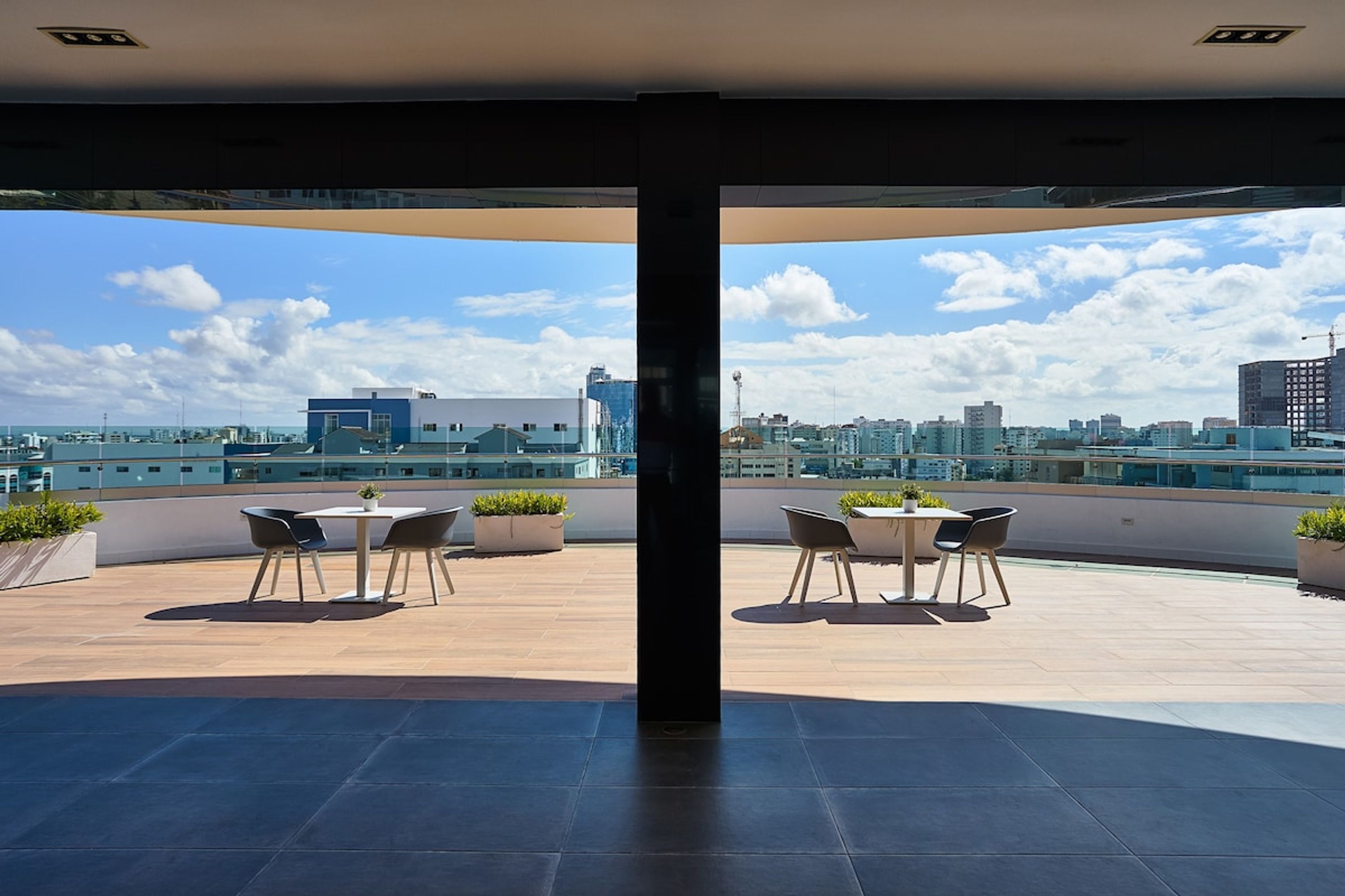 A rooftop terrace with two tables and chairs, potted plants, and a city skyline view.