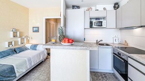Een ruimtelijk ingerichte leefruimte met een kitchenette, een bed met een blauw-wit gestreept kussen en een klein eethoekje.