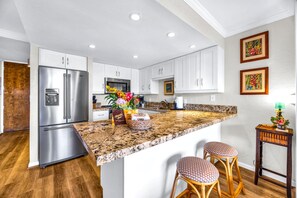 A kitchen with white cabinets, a stainless steel refrigerator, a granite countertop, and two wicker bar stools. - Papakea Villas | Ocean View Studio | Car Incl. w/ 6+ Nights | PKD-303 by KBM (Lahaina)