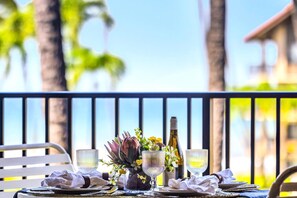 A dining table set with plates, glasses, and a bottle, with a view of palm trees and a building in the background. - Papakea Villas | Ocean View Studio | Car Incl. w/ 6+ Nights | PKD-303 by KBM (Lahaina)