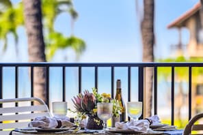 A dining table set with plates, glasses, a bottle, and a vase with flowers, overlooking a balcony with a railing and palm trees in the background. - Papakea Villas | Ocean View Studio Sleeps 4 | Car Incl. w/ 6+ Nights | PKD-303 by KBM (Lahaina)