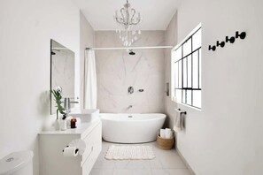 Une salle de bain moderne avec une baignoire sur pieds, un grand miroir, un lustre et une fenêtre avec des stores.