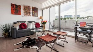 A modern living room with a sofa, coffee table, and leather poufs, a glass dining table, and a large window with a city view.