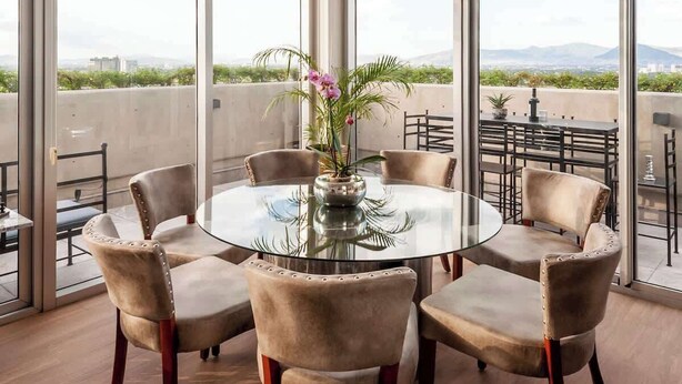 A round glass table with six chairs, a vase with flowers, and a city view through large windows.