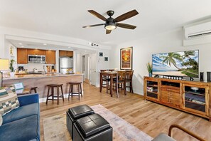A modern kitchen with wooden cabinets, a dining area with a wooden table and chairs, a living room with a blue sofa, a black leather ottoman, and a wooden entertainment center with a TV. - Papakea Villas | Ocean View 2 BR Sleeps 8 | Car Incl. w/ 6+ Nights | PKD ML-3523 by KBM (Lahaina)