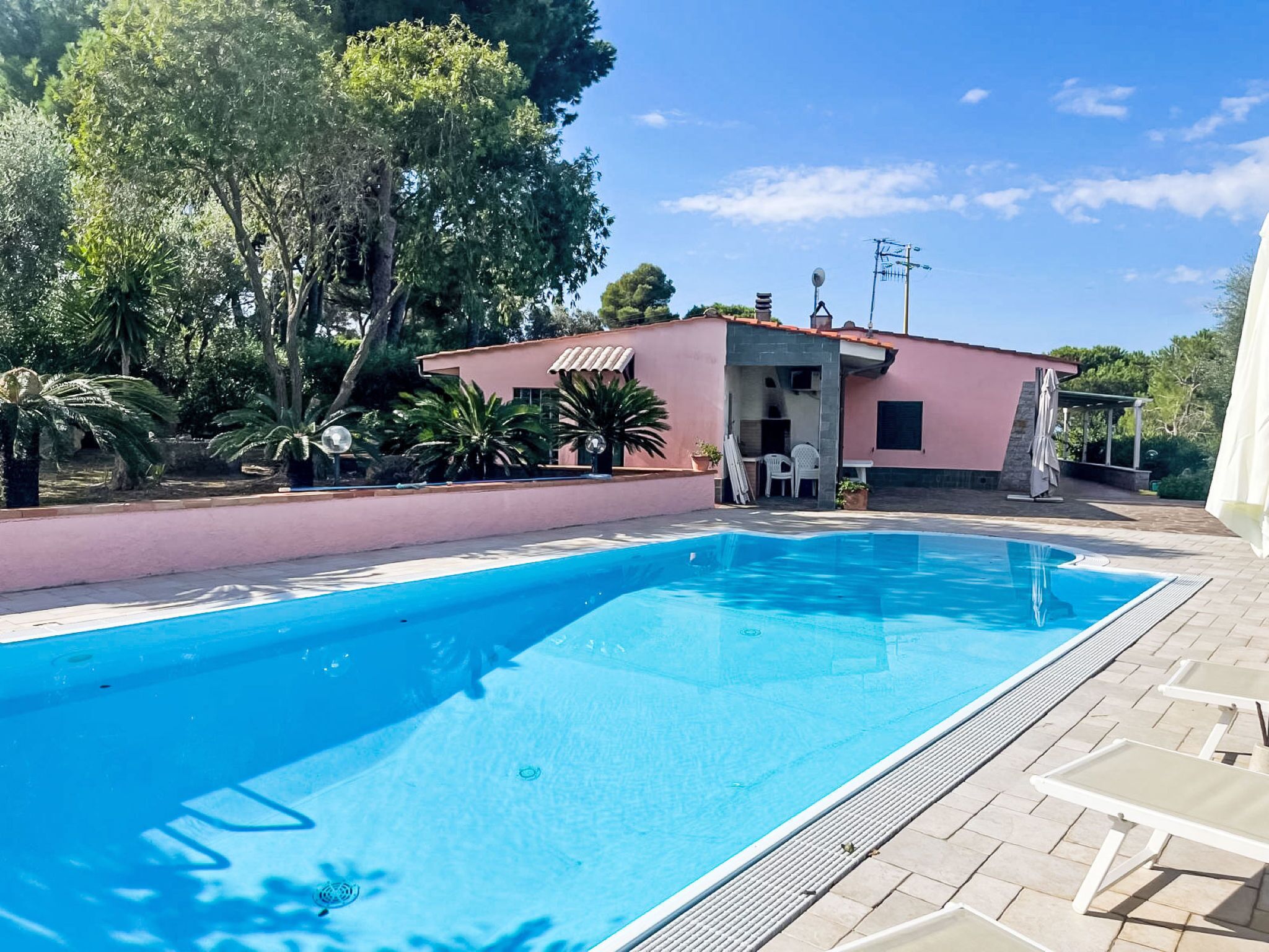 Une piscine avec une terrasse carrelée, un bâtiment à la façade rose et une végétation luxuriante tout autour.