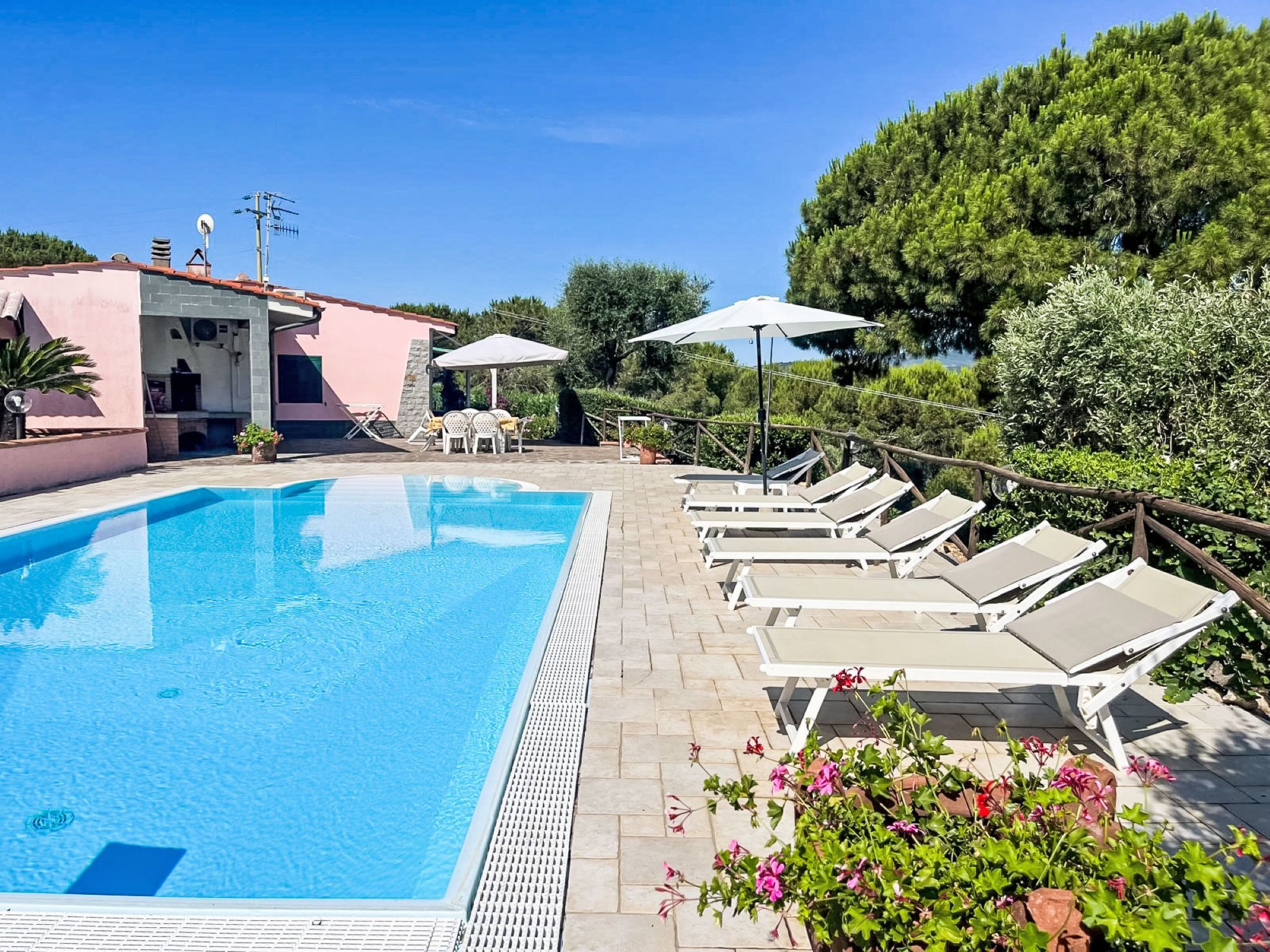 Une piscine entourée de verdure, agrémentée de chaises longues et de parasols, et bordée d’un bâtiment à la façade rose.