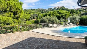 Un espace piscine doté de chaises longues, d’une terrasse en pierre et offrant une vue sur une colline parsemée d’arbres et de bâtiments.