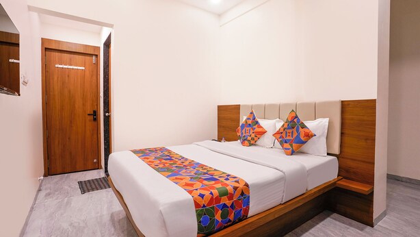 A hotel room with a bed, a wooden headboard, a door, and a small bedside table. - Fabhotel Vishwas (Chhatrapati Sambhajinagar)