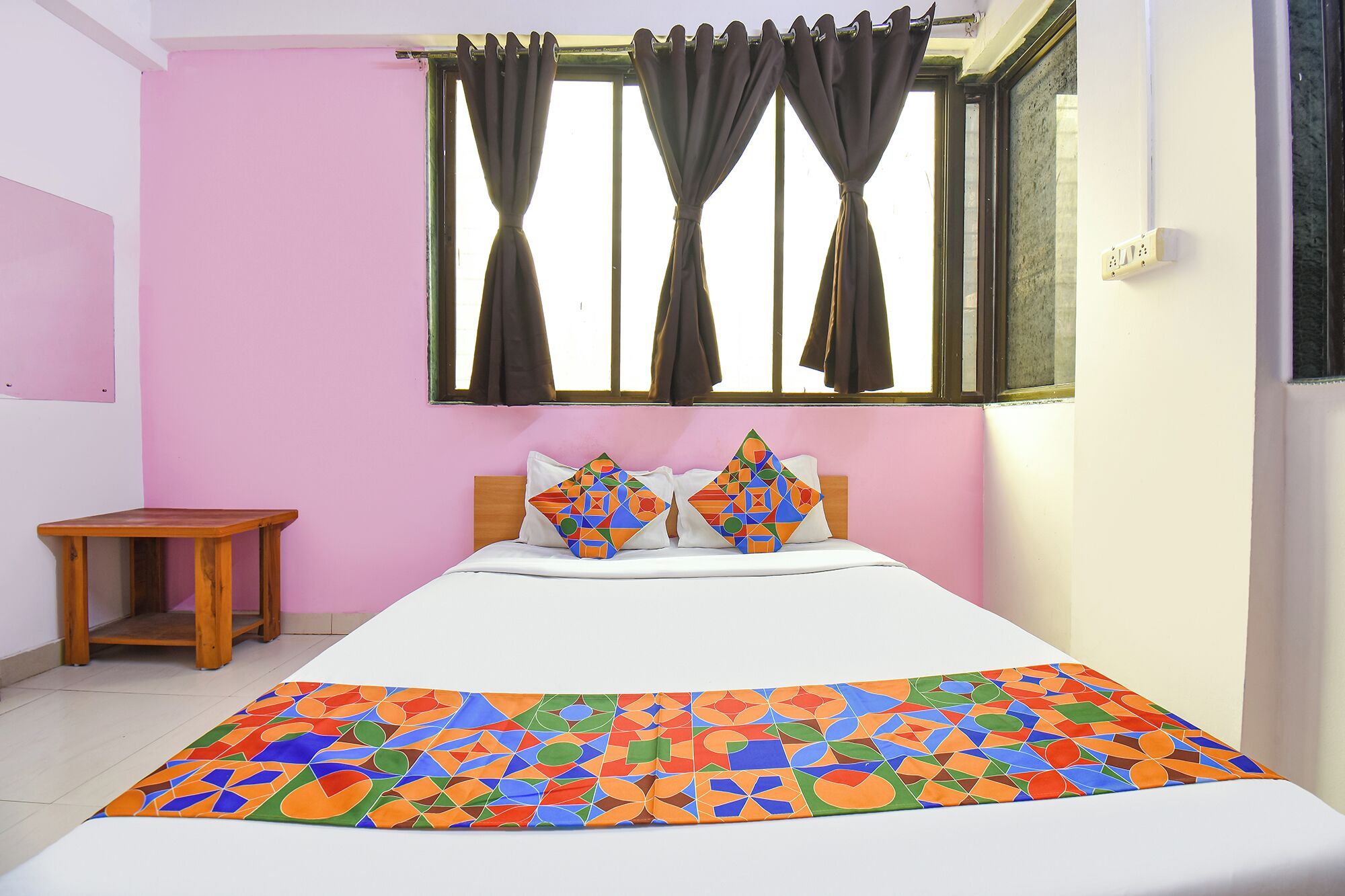 Un dormitorio con cama, almohadas, una mesita y una ventana con cortinas.