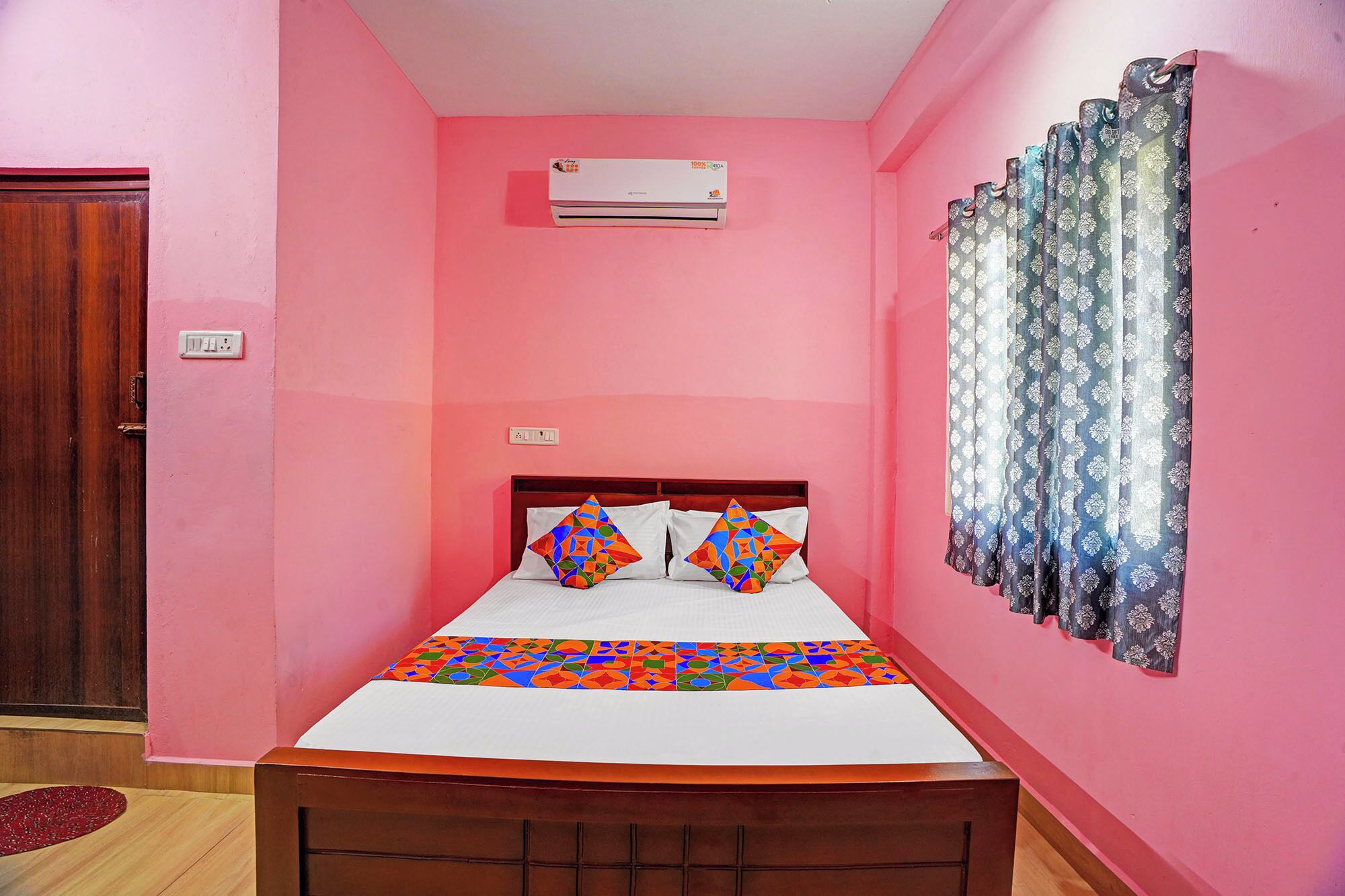 A room with pink walls, a bed with colorful geometric pillows, a wooden headboard, a window with patterned curtains, and an air conditioning unit on the wall.