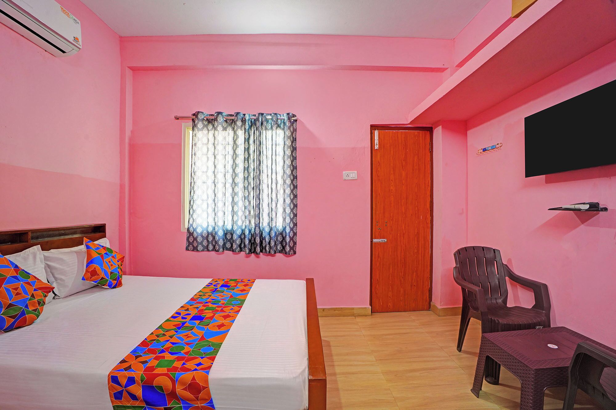 A pink-themed room with a bed, a chair, a small table, a TV, and a window with curtains.