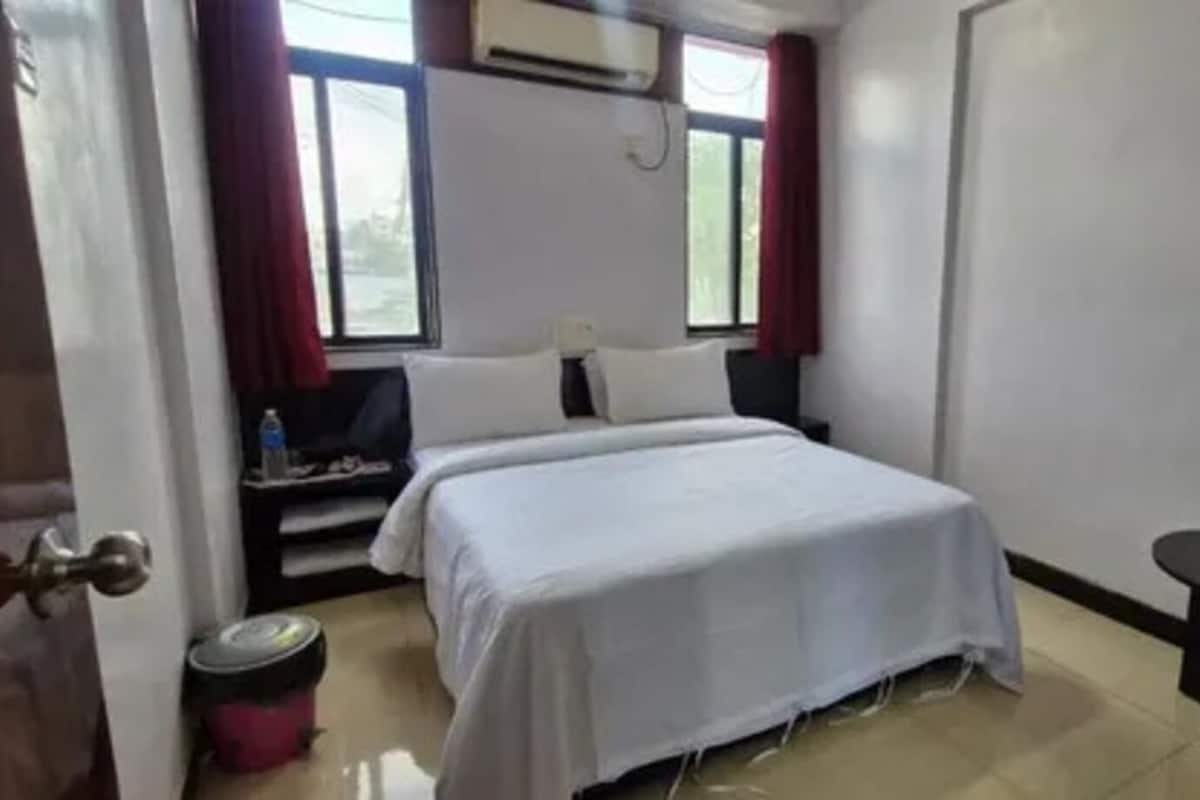 A hotel room with a bed, a small table, a trash bin, and a window with curtains.