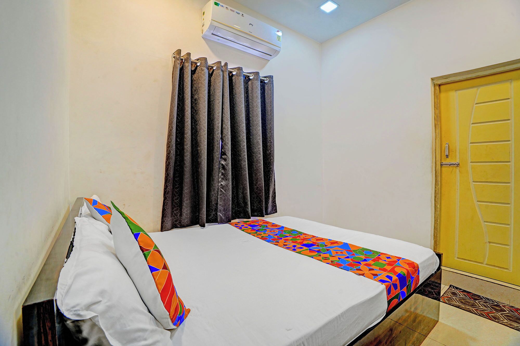 A single bed with a colorful geometric-patterned bedspread, a white pillow, and a dark patterned curtain. An air conditioning unit is mounted on the wall above the bed.
