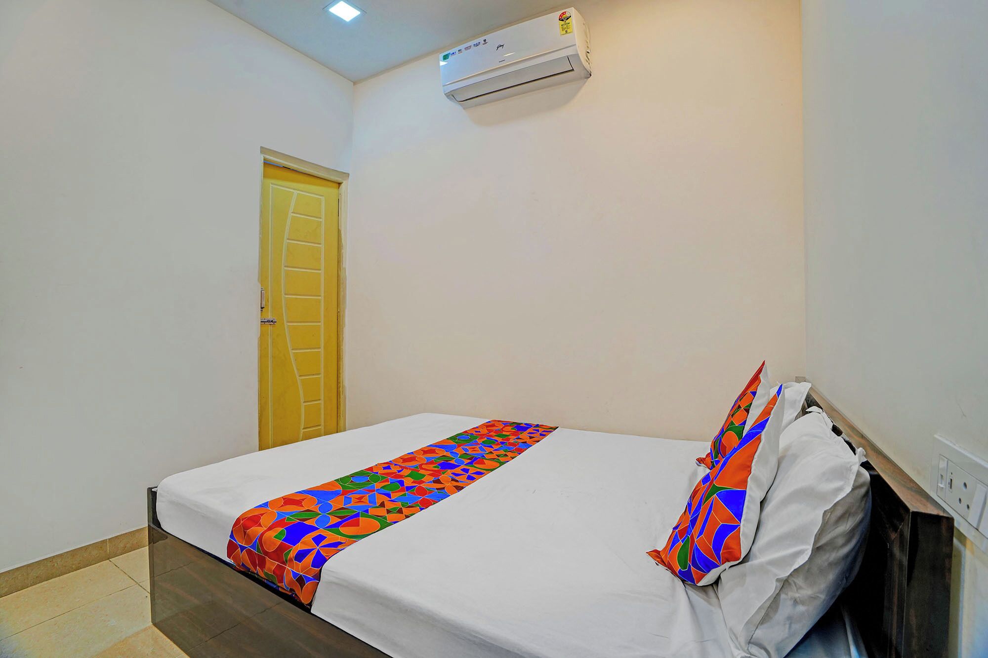 A single bed with a colorful geometric-patterned bedspread and pillows, a plain white wall, a yellow door, and a ceiling-mounted air conditioner.