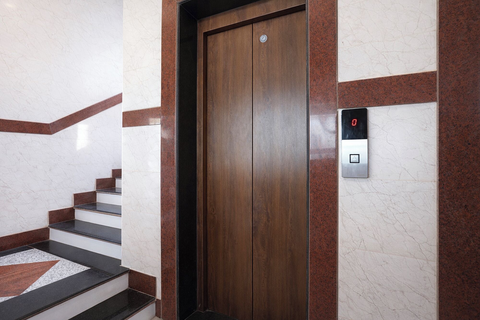 Puerta de ascensor de madera con pantalla digital, paredes de mármol y una escalera con peldaños blancos y negros.