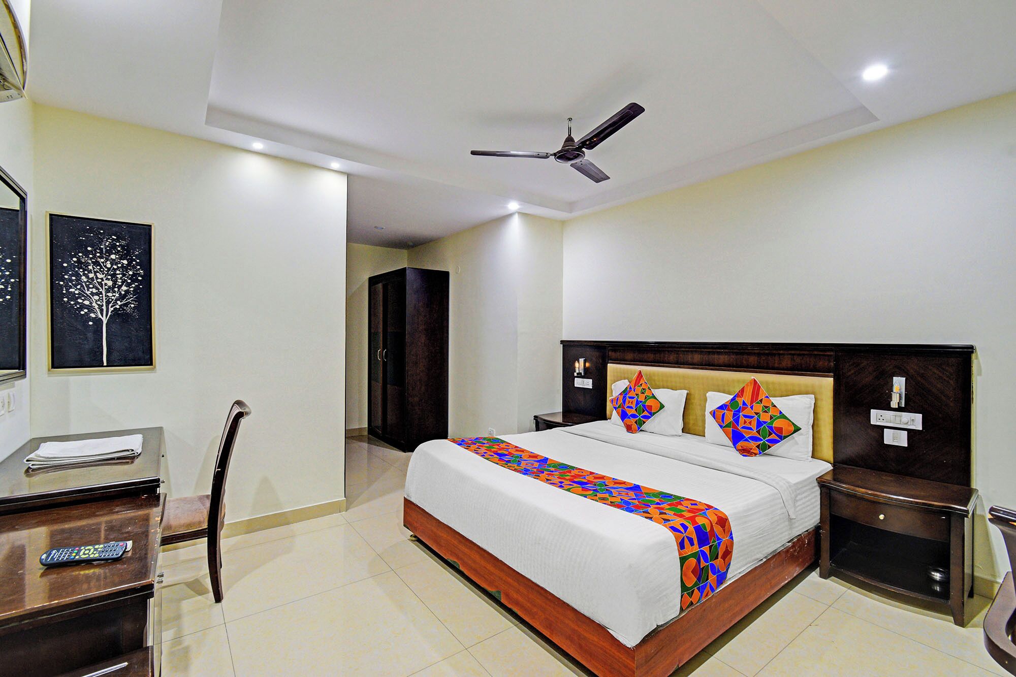 A hotel room with a bed, bedside tables, a ceiling fan, a wardrobe, and a desk with a chair.