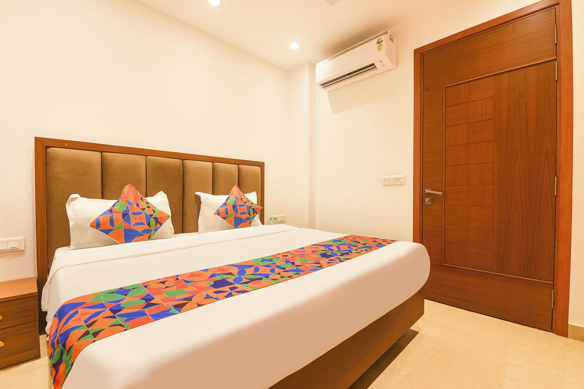 A hotel room with a bed, a wooden door, an air conditioning unit, and a nightstand.