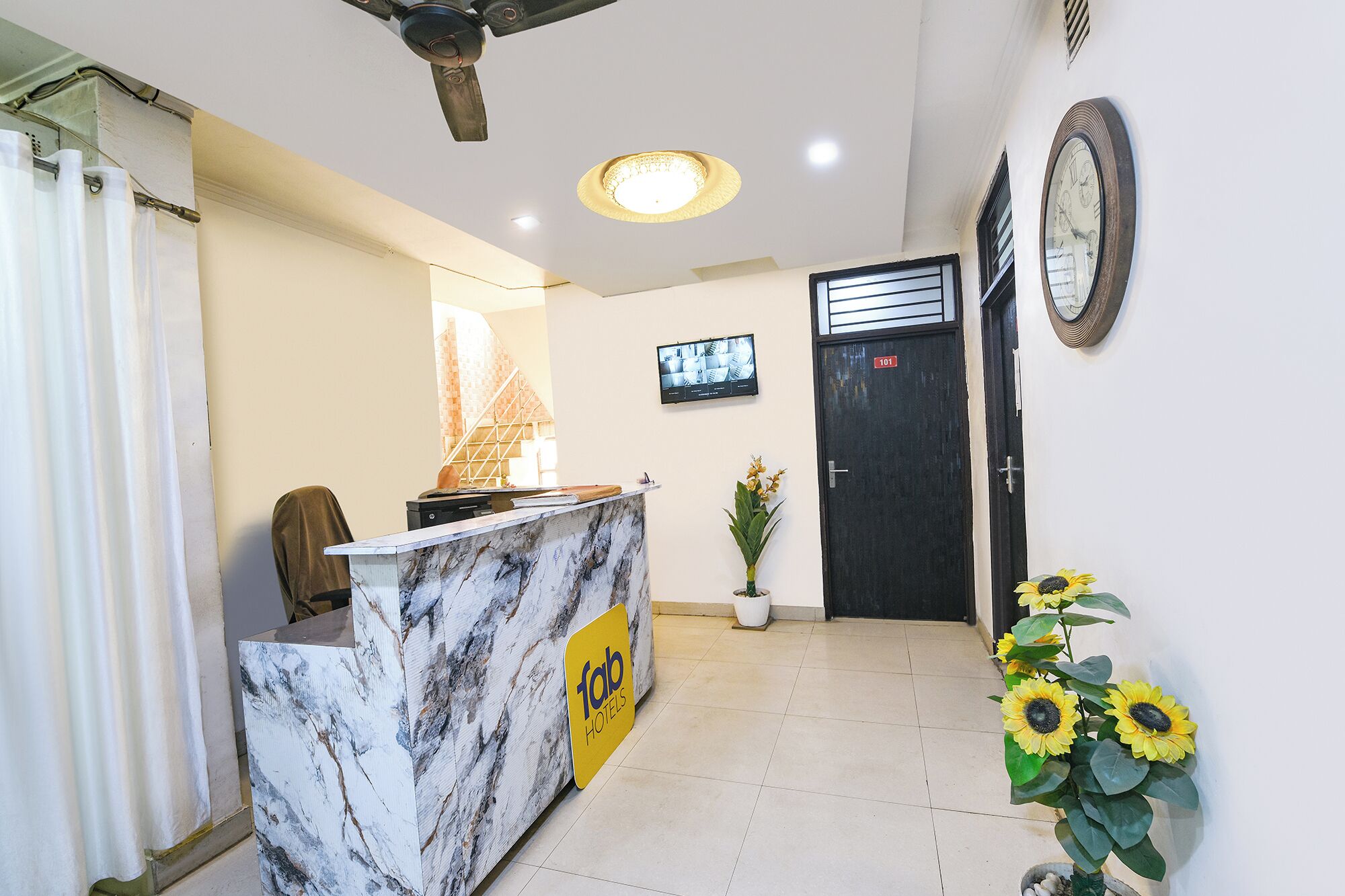 A reception desk with a marble top, a ceiling fan, a television on the wall, a door with a number, and a potted plant with sunflowers.