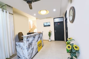 A reception desk with a marble top, a ceiling fan, a television on the wall, a door with a number, and a potted plant with sunflowers. - Fabhotel Mb Residency (New Delhi)