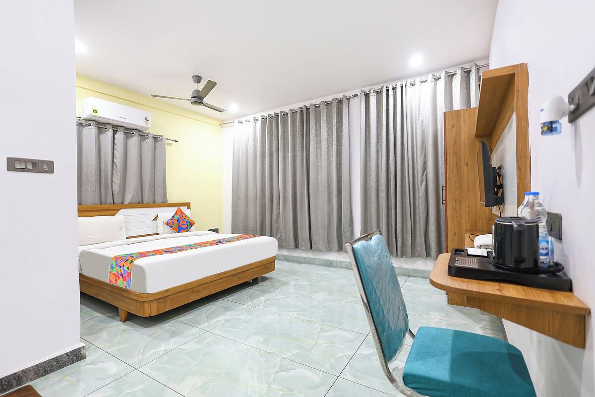 Une chambre d’hôtel comprenant un lit, un bureau avec une chaise, un miroir, un ventilateur de plafond et une fenêtre avec des rideaux.